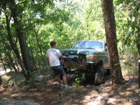 1978 Ford Bronco - Man pulls on winch cable attached to a modified green truck stuck in the woods.