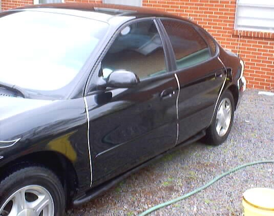 Black 1999 Ford Taurus sedan, three-quarter front view, parked on gravel near a brick building