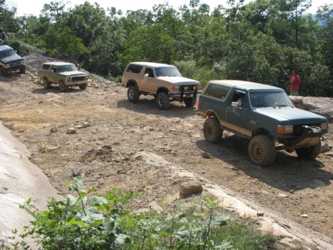 1978 Ford Bronco - Several off-road vehicles, including a green Ford Bronco, navigate a rocky, wooded trail.