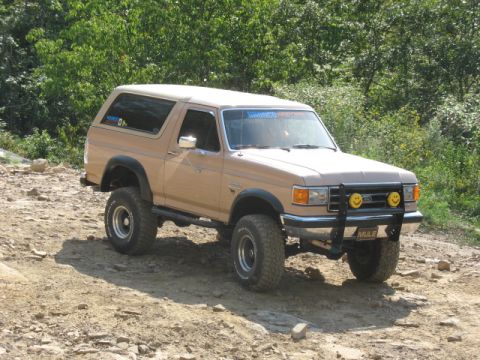 1978 Ford Bronco - A tan Ford Bronco is parked on a dirt trail with trees in the background.