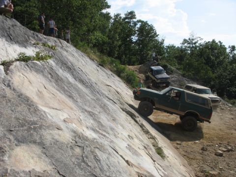 1978 Ford Bronco - Several off-road vehicles navigate a steep, rocky incline on a sunny day with trees in the background.