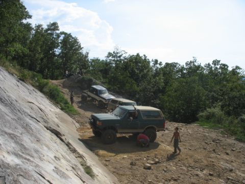 1978 Ford Bronco - Multiple off-road vehicles navigate a steep, rocky incline surrounded by dense trees.