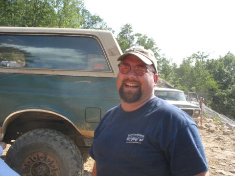 1978 Ford Bronco - A man smiles next to a 1978 Ford Bronco SUV, with another vehicle visible in the background on a rocky, wooded terrain.