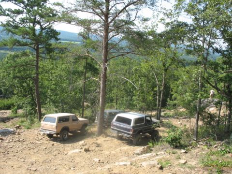 1978 Ford Bronco - Two Ford Broncos navigate a wooded off-road trail with a scenic overlook in the background.