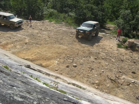 1978 Ford Bronco - Two SUVs navigate a rocky, off-road trail with wooded hills in the background.