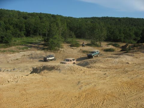 1978 Ford Bronco - Multiple SUVs navigate a challenging dirt terrain under a clear blue sky, with a dense forest bordering the area.