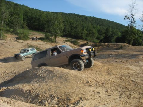 1978 Ford Bronco - Two Ford Broncos navigate an off-road course with wooded hills in the background.