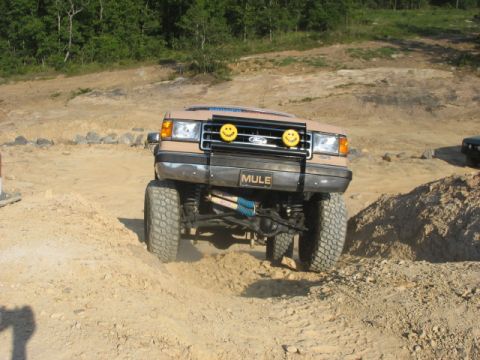 Tan Ford Bronco with "MULE" license plate and smiley face lights on front grille, driving on dirt terrain