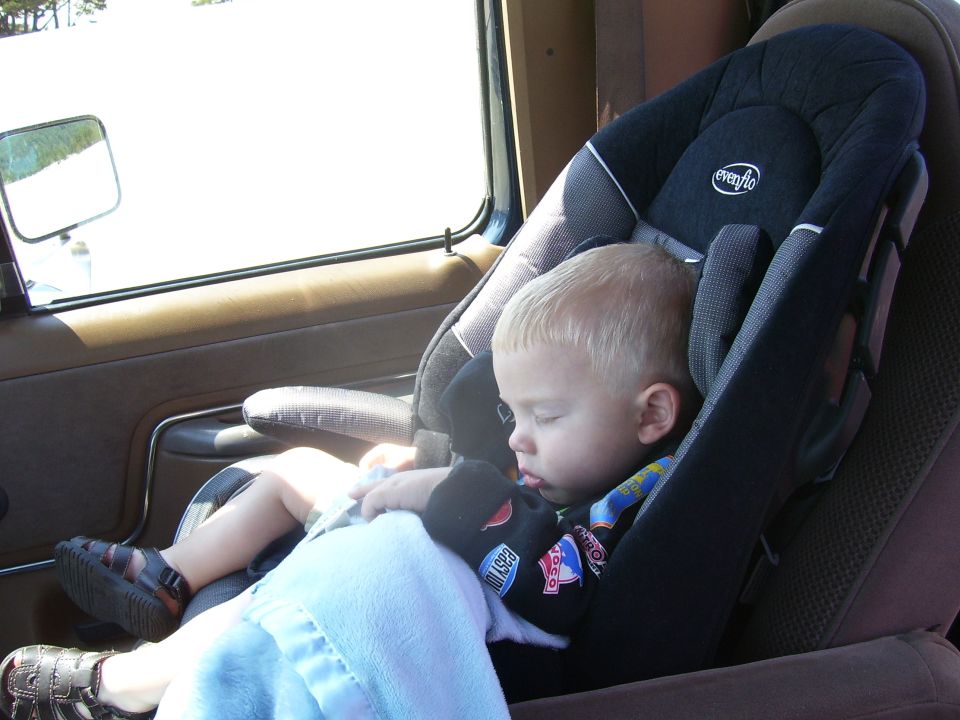 Child sleeping in a car seat inside a vehicle, with a side mirror visible through the window