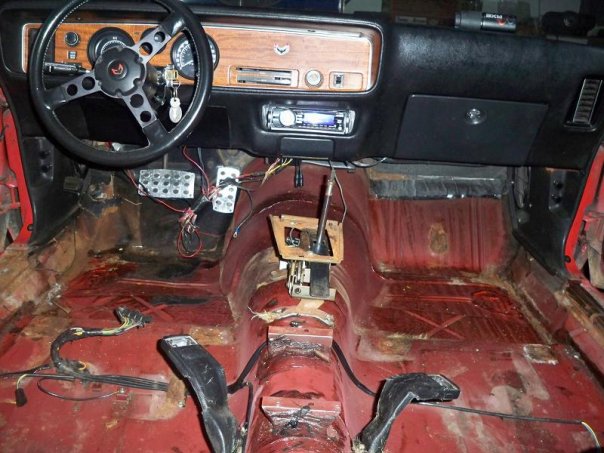 Car interior with red floor, black dashboard, and steering wheel. The floor is stripped and shows rust