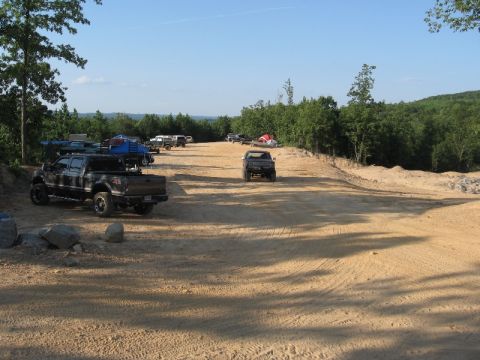 1978 Ford Bronco - Multiple trucks are gathered in an outdoor dirt lot with a wooded area and hills visible in the distance.