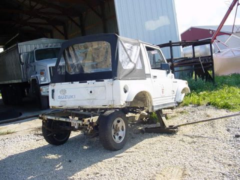 1992 Ford Bronco - Suzuki Samurai with soft top is jacked up, with a large truck visible in the background.