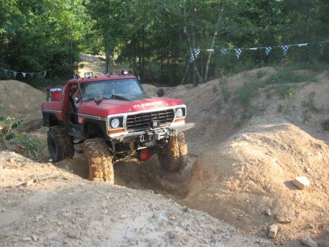 1978 Ford Bronco - A red pickup truck with off-road modifications drives through a challenging dirt terrain.