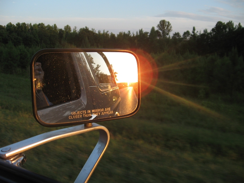Side mirror reflection shows sunset over a road; 'Objects in mirror are closer than they appear' text visible