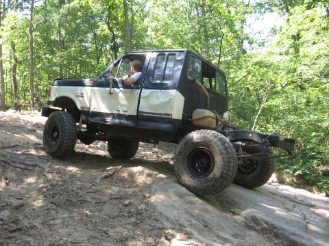 1978 Ford Bronco - Off-road truck navigating rocky terrain in a forest setting. Modifications include large tires and a custom body.