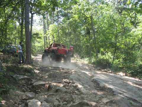 1978 Ford Bronco - A red 1978 Ford Bronco navigates a challenging, rocky off-road course surrounded by dense trees.