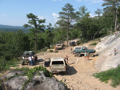 1978 Ford Bronco - Several off-road vehicles are gathered on a rocky, wooded hillside under a clear blue sky.