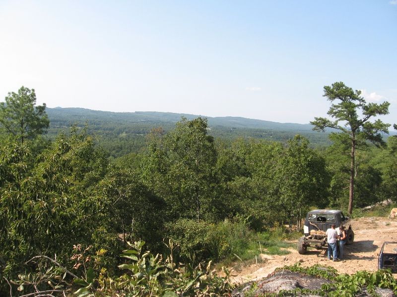 Off-road vehicle parked on dirt with people standing nearby, overlooking a wooded valley