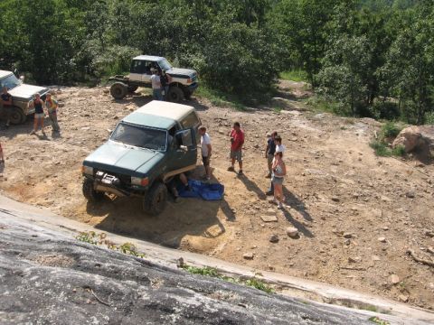 1978 Ford Bronco - Several people are gathered around a green SUV with its doors open, parked on a rough dirt path surrounded by trees.