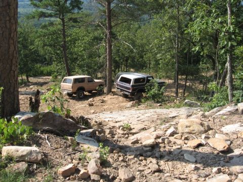 1978 Ford Bronco - Two classic Ford Bronco SUVs navigate a challenging off-road trail surrounded by dense forest and rocky terrain.