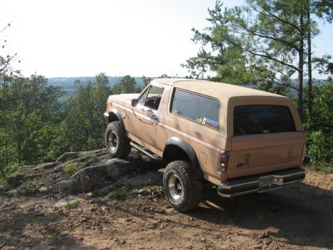 1978 Ford Bronco - A tan Ford Bronco navigates a rocky, off-road path with trees and a distant landscape in the background.