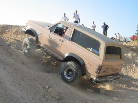1978 Ford Bronco - Tan/desert-sand Ford Bronco climbing a steep dirt bank at an off-road park, shot from the rear three-quarter as the driver works up the gra…