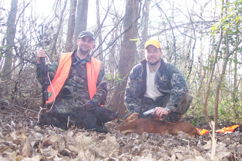 Two guys in camouflage and orange vests pose with a large hog and a smaller piglet in a wooded area