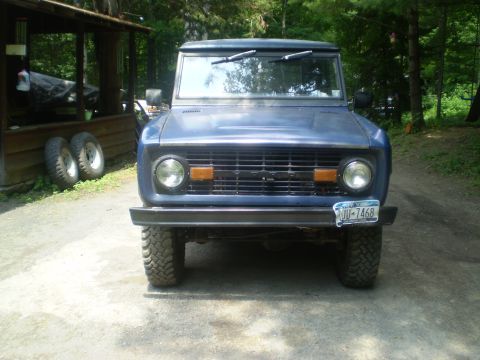 1979 Ford Bronco - This blue Ford Bronco is parked outside a wooden structure with spare tires nearby.