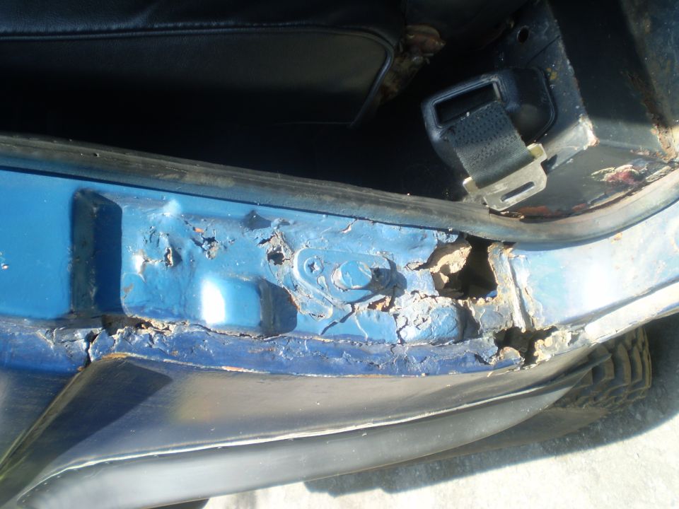 Close-up of severe rust damage on a blue vehicle's door sill, with a black seatbelt visible