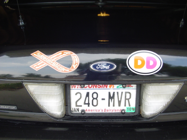 Rear of a 2000 Ford Crown Victoria showing the trunk lid with a 'Support Bacon' ribbon sticker, a Ford badge, a Dunkin' Donuts sticker, and a Wisconsin license plate reading 248-MVR