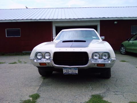1972 Ford Ranchero - A white pickup truck with a distinctive grille and hood scoop is parked outside a red building.