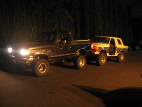 1989 Ford Bronco - A dark pickup truck is parked next to a white and blue SUV at night.