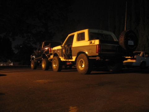 1989 Ford Bronco - Two modified Ford Broncos are parked side-by-side at night, with one a spare tire mounted on its rear.