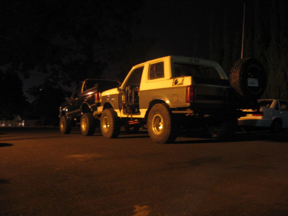 Two lifted Ford Broncos parked at night. One is white and gray with a spare tire on the back. The other is dark colored