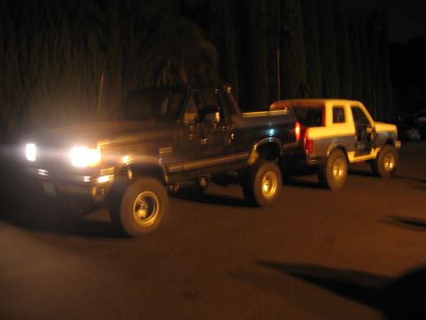 1989 Ford Bronco - Two SUVs are parked on a road at night, with the front vehicle's headlights illuminated.