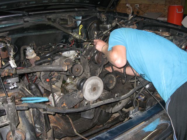 Person in blue shirt leaning into open engine bay working on engine with visible fan and belts