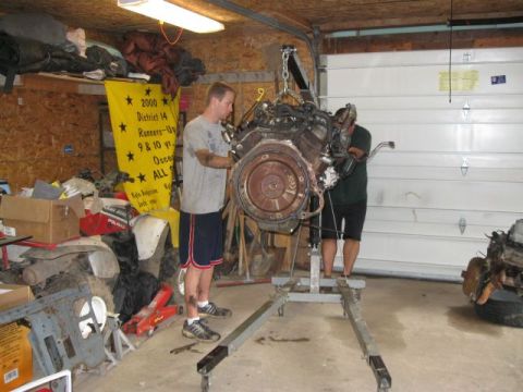 1992 Ford Bronco - Two men work on an engine suspended by a hoist in a cluttered garage.