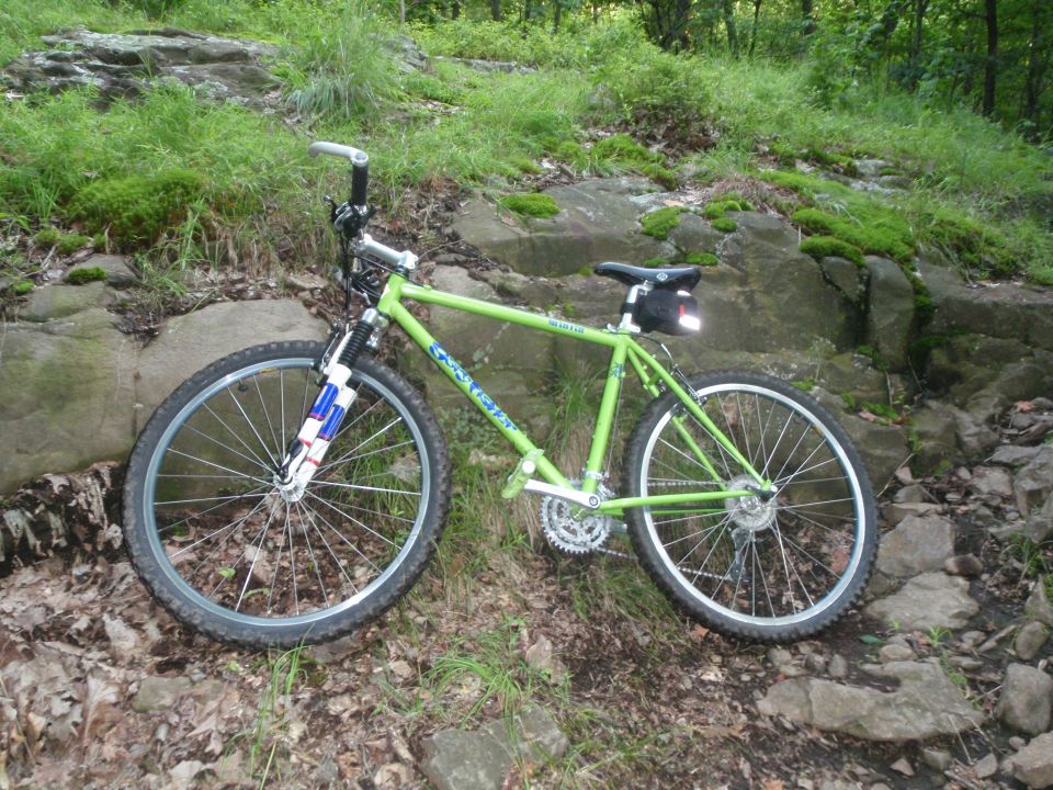 Green mountain bike parked on a rocky, wooded trail. The bike has a suspension fork and a saddle bag