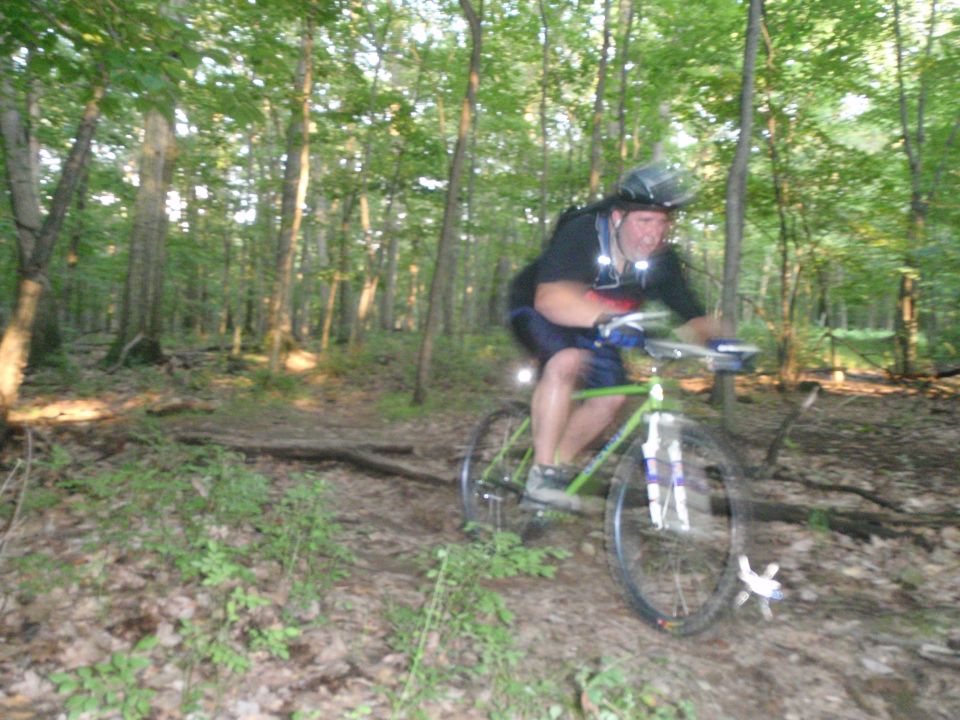 Guy on a green bike riding down a wooded trail. He's wearing a helmet and backpack