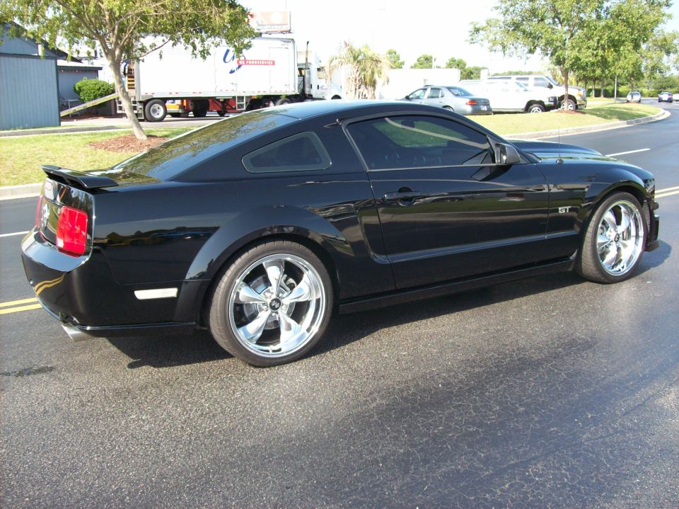 Black Ford Mustang GT coupe with chrome five-spoke wheels, rear three-quarter view in a parking lot