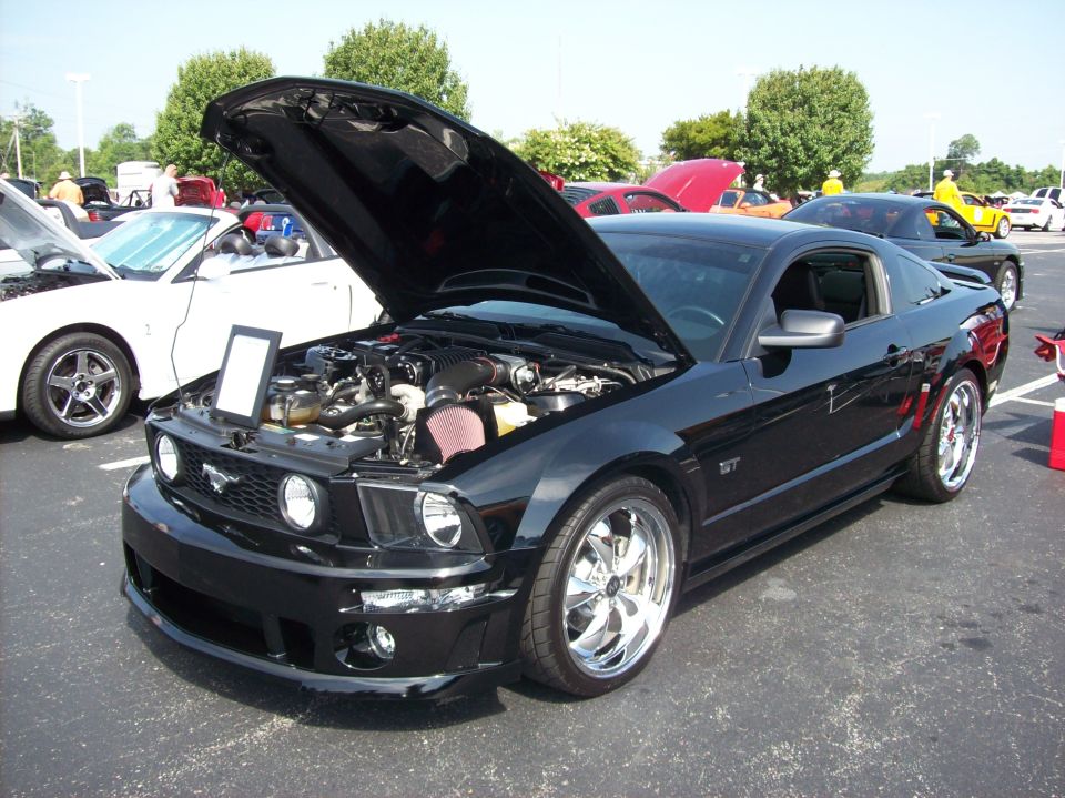 Black 2007 Ford Mustang GT with hood open, showing engine and aftermarket wheels. White Mustang convertible parked to the left