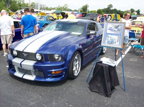 2007 Ford Mustang - A blue 2007 Ford Mustang with white racing stripes is displayed at an outdoor event, next to a framed photo of the car.