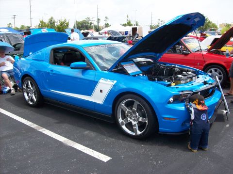 2007 Ford Mustang - A child inspects the engine of a blue Ford Mustang at a car show. Another vehicle is visible in the background.