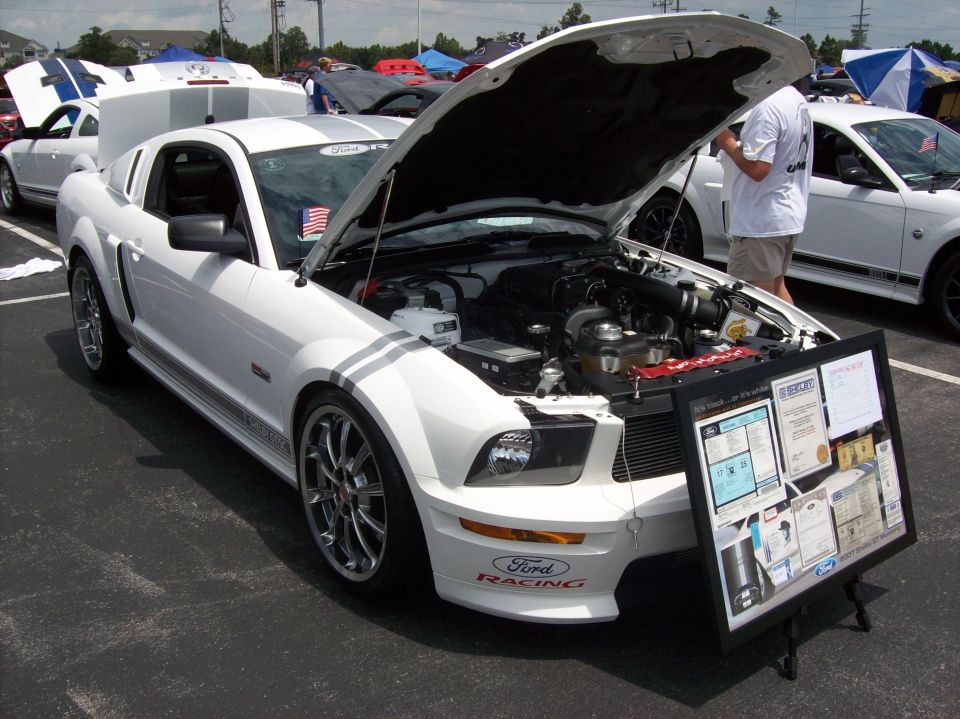 White Ford Mustang with racing stripes, hood open revealing engine, and a display board in front