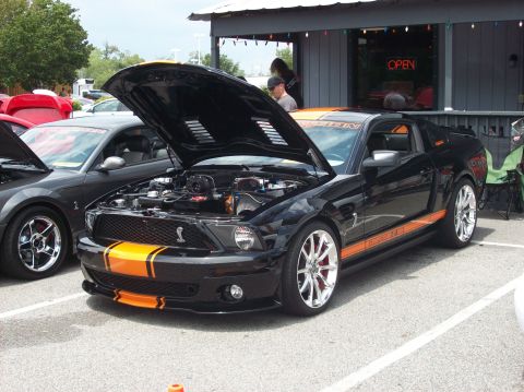 2007 Ford Mustang - A black 2007 Ford Mustang with orange racing stripes and its hood open is parked next to another car.