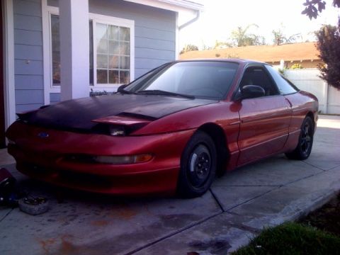 1995 Ford Probe - A red coupe with a black hood is parked on a concrete driveway in front of a house.