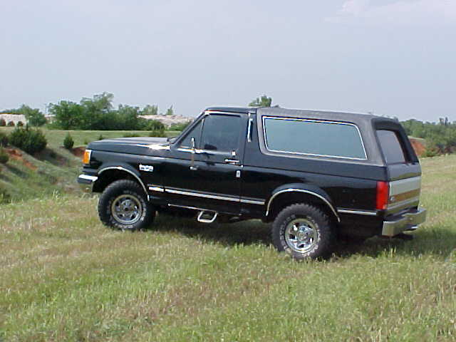 Black Ford Bronco SUV parked on grassy hill, 3/4 front view. Chrome wheels, aftermarket step bars visible