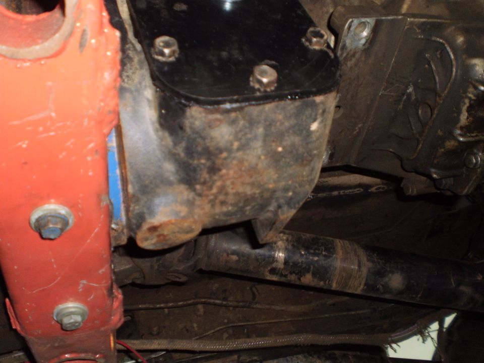 Rusty bolted skid plate next to red crossmember with driveshaft underneath