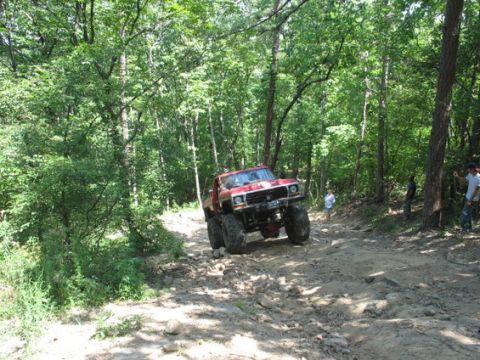 1979 Ford Bronco - A modified red Ford F-150 navigates a challenging off-road trail surrounded by dense trees.