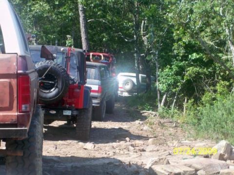 1979 Ford Bronco - A line of off-road vehicles, including a 1979 Ford Bronco, navigate a rocky trail through a wooded area.